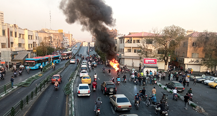 Ett fordon brinner på en stor gata i Iran. Folk och bilar är samlade runt den.