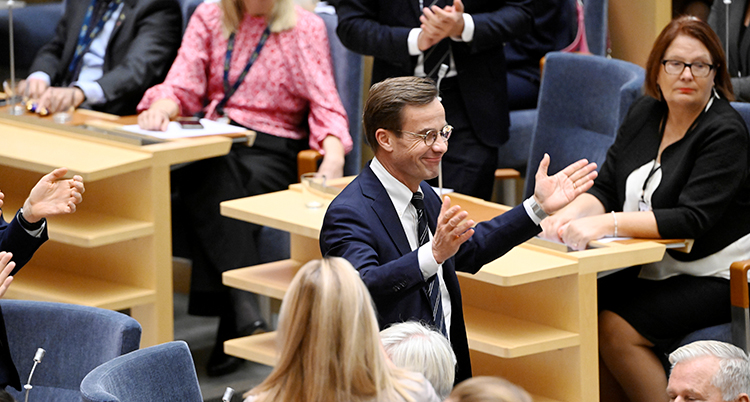 Ulf Kristersson står upp i riksdagen och ser glad ut.