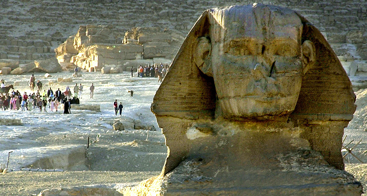 Ett stort huvud i sandsten syns i förgrunden. Bakom går människor och tittar.