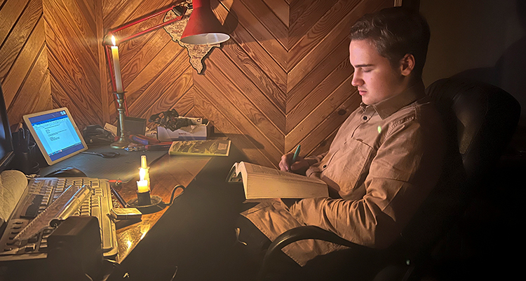 En pojke sitter och läser med tända stearinljus. Han har också en dator framför sig.