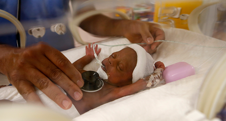 En liten baby ligger i en kuvös. Babyn har en mössa. En slang går in i babyns näsa. En vuxen hand håller ett stetoskop mot babyns mage.