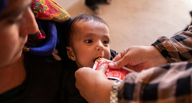 En baby ligger i famnen på en kvinna. Händer sträcker fram en påse med mat mot barnets mun.