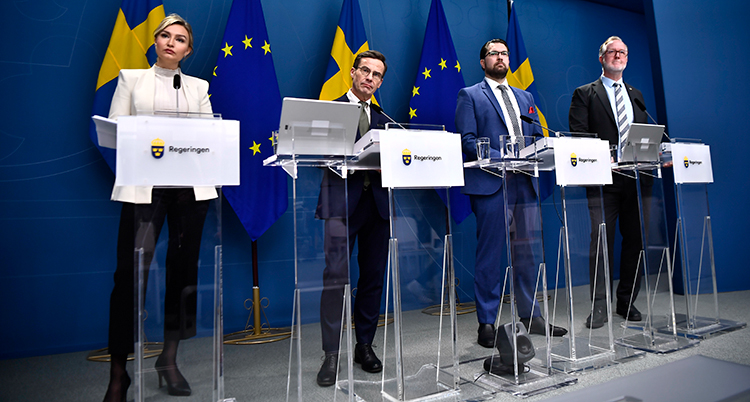 De fyra står bakom varsin talarstol i glas. Bakom syns svenska flaggor och EU-flaggor