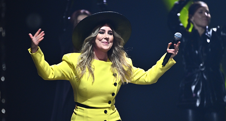 Hon har en gul dräkt och en svart hatt. Hon håller i en mikrofon och gör gester med armarna.