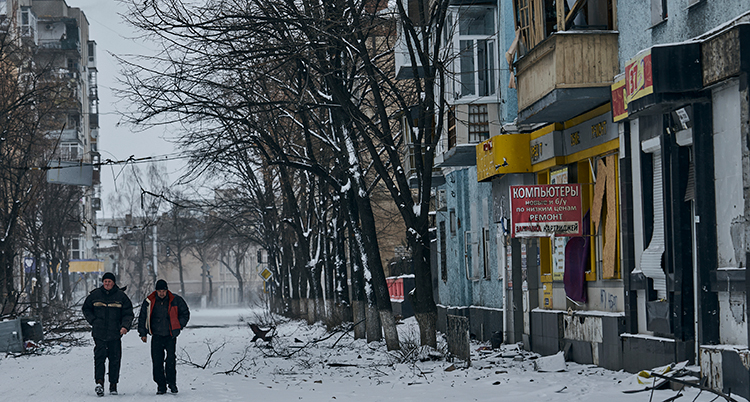 Två män går på en snöig gata i staden. Husen runt dem ser sargade ut.