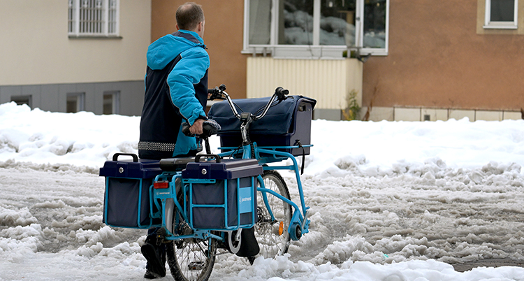 En brevbärare drar en cykel med postväskor genom snön på en väg. Ett hyreshus syns bakom