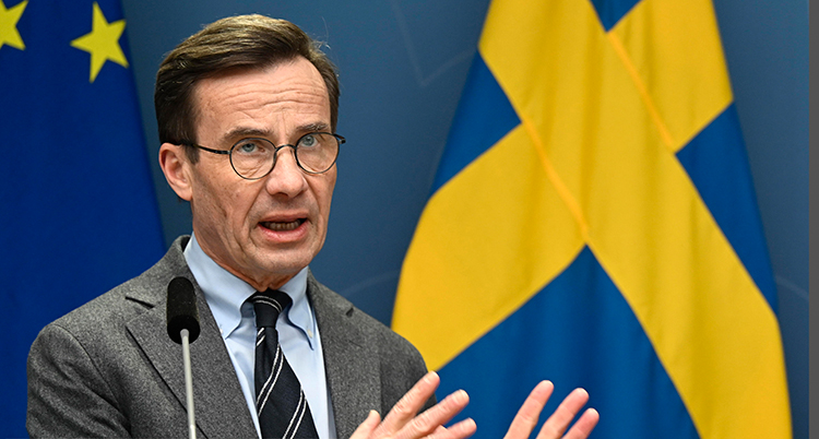 han har glasögon, kostym och slips. Han står framför en svensk flagga och pratar i en mikrofon. Han ser allvarlig ut.