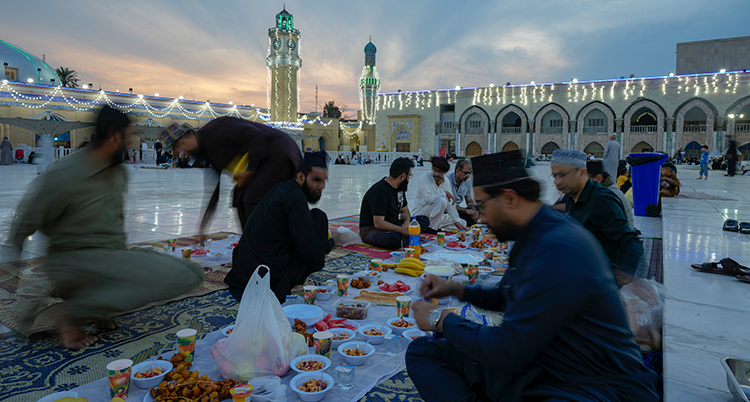 Flera män sitter på marken längs en duk på marken. Det står mat på duken. Bakom syns en moské.