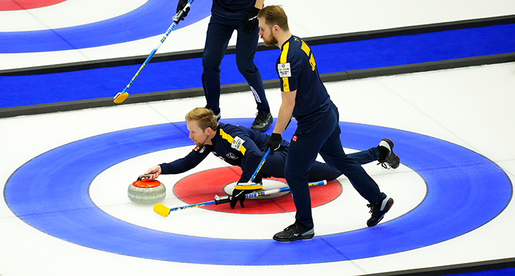 Spelare på en plan för curling. En spelare ligger ner på isen och ska skjutsa iväg en sten. Två spelare står bredvid med sopkvastar i händerna.