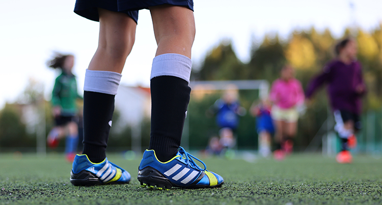 Ett barn med fotbollskläder på en fotbollsplan. Bilden visar bara barnets fötter och ben.