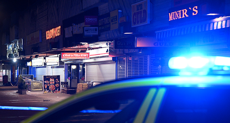 Det är kväll på bilden. En del av en polisbil syns i förgrunden. En upplyst ingång syns i bakgrunden och skyltar på butiker och restauranger.