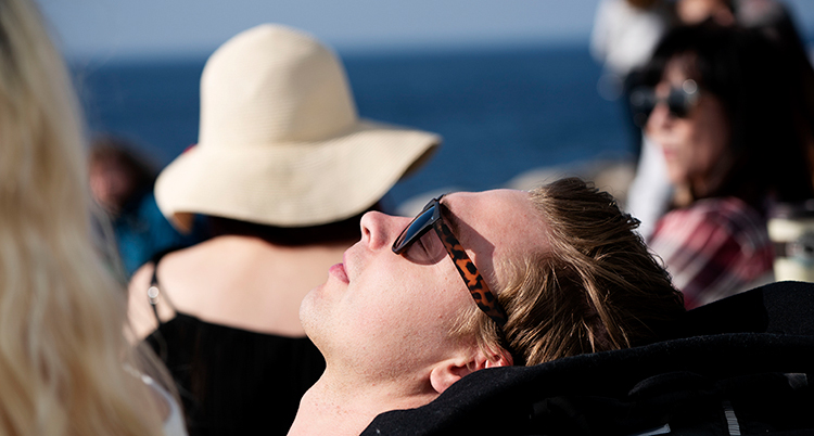 Ett ansikte med solglasögon solar. I bakgrunden syns en person i hatt.