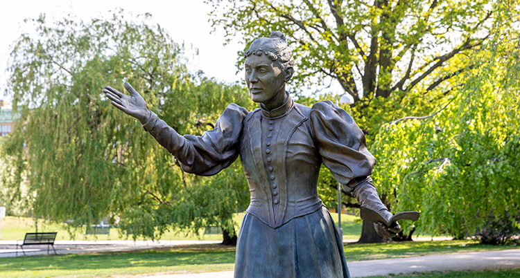 En staty av en kvinna står i en park med träd med ljusgröna blad.