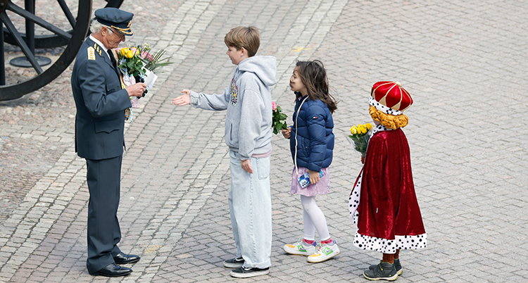 Tre barn står på rad för att ge kungen blommor. Pojken längst fram sträcker fram handen för att hälsa på kungen. Sist står ett barn som klätt ut sig till kung med krona och mantel.
