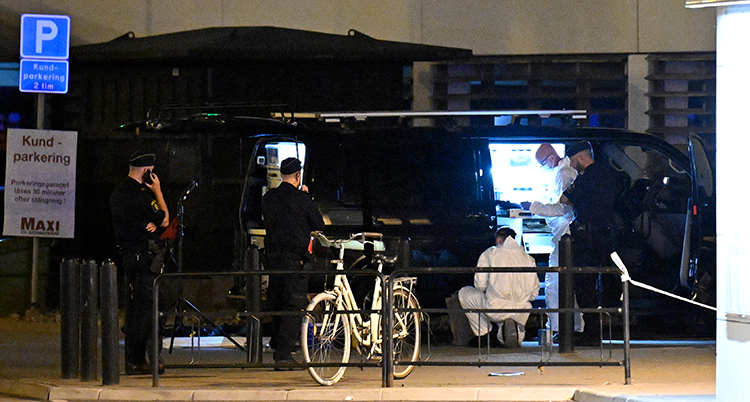 Personer syns vid en mörk parkeringsplats. En skylt syns framme till vänster. En cykel står mitt i bilden.