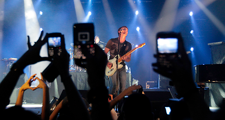 En kille står på en scen och sjunger och spelar gitarr. I publiken är det flera som filmar med sina mobiler.