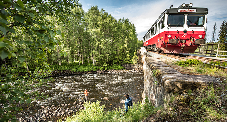 Ett rött tåg kör över en å där två personer fiskar.