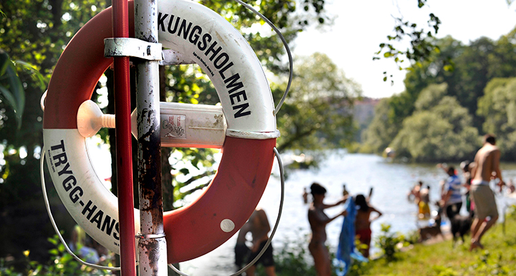 Livboj i förgrunden. Badande personer vid sjö i bakgrunden.