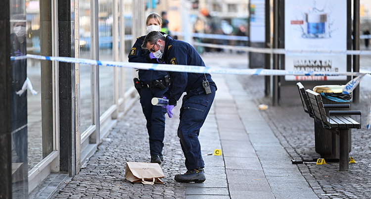 Två poliser med uniformer och munskydd jobbar på gatan. En papperspåse ligger på marken framför dem. Det är stora fönster på vänster sida. Avspärrningsband syns i förgrunden.