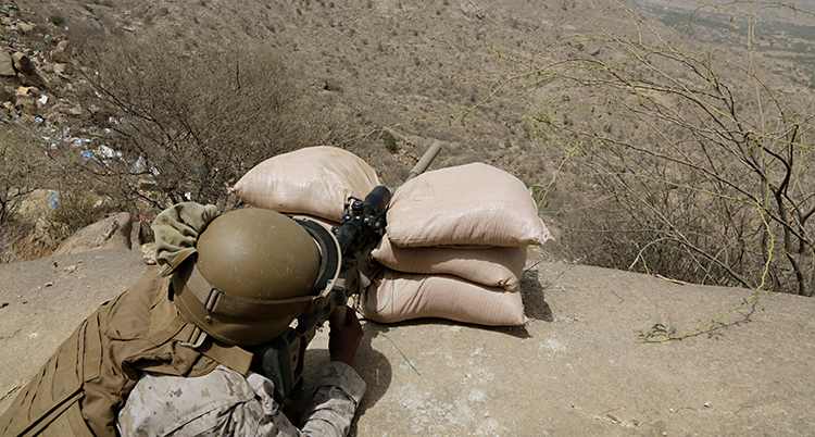 En soldat ligger och siktar med sitt gevär ner mot en dalgång.