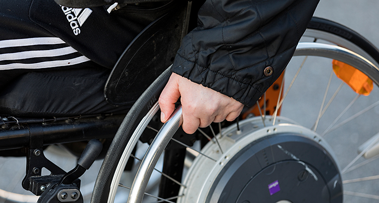 En person som sitter i en rullstol. Närbild på ena handen och hjulet.