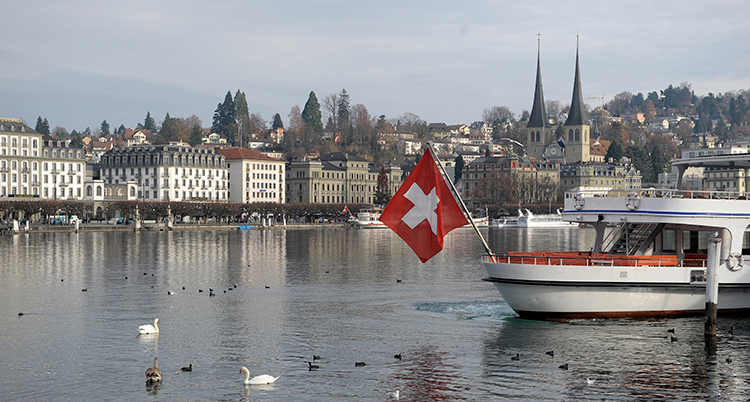 En båt med en schweizisk flagga i aktern syns till höger i bilden. Till vänster syns vatten och hus vid strandkanten.
