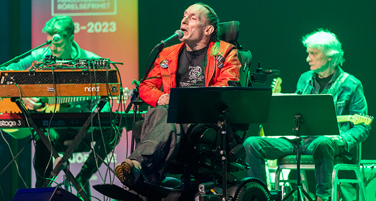Han sitter i en rullstol på en scen. Han sjunger i en mikrofon. Bakom syns två musiker.