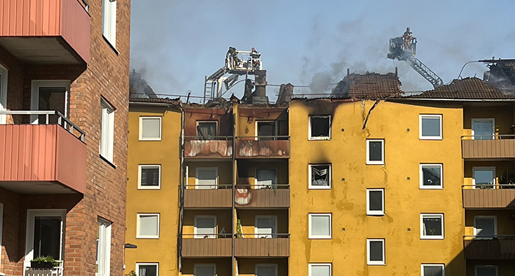 Personer jobbar vid taket på ett stort hus med lägenheter. Det är skador på huset.