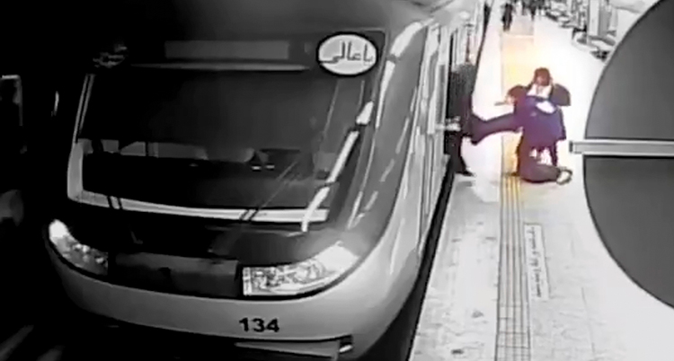 en suddig övervakningsbild visar ett tåg vid en perrong. en person på perrongen ser ut att hålla fast en annan person.