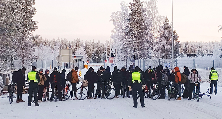 En stor grupp med människor står med cyklar på en snöig väg. Bakom syns en gränsstation.