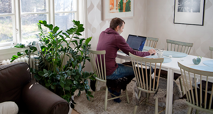 En man sitter vid ett bord i vardagsrummet. Han sitter och jobbar med en dator.