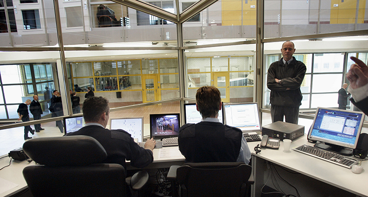 Folk sitter vid ett skrivbord med skärmar och tittar på fängelset genom en glasruta.