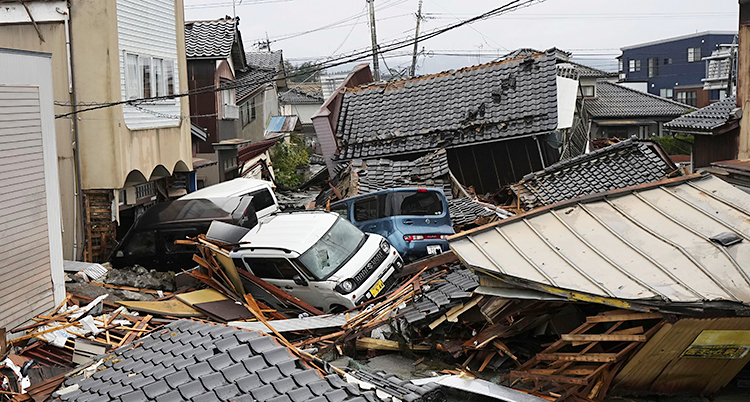 En stad i Japan. Det är en stor röra. Flera hus har rasat ihop och några bilar ligger mitt i röran.