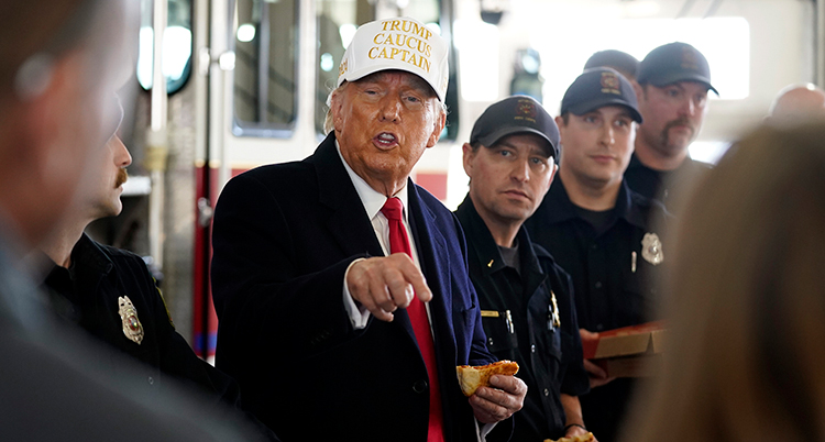 Trump med en pizza i handen pekar på något med andra handen. Det står flera män runt honom och lyssnar.