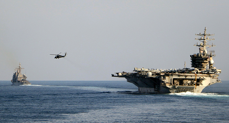 Ett stort krigsfartyg är på havet. Ett mindre fartyg syns en bit bort och en helikopter flyger i luften.