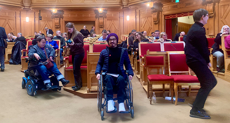Han sitter i en rullstol i en sal i riksdagen.