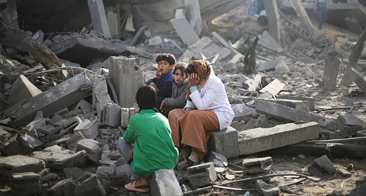 En kvinna och flera barn sitter vid ett utbombat hus. KVinnan ser ledsen ut.