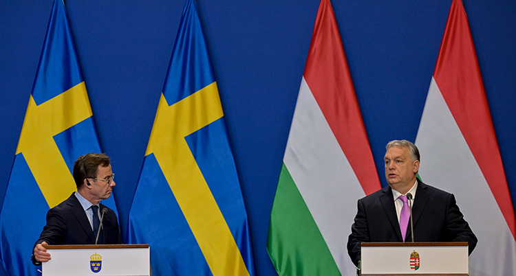 De står i varsin talarstol. Ulf Kristersson tittar mot Viktor Orbán. Bakom dem syns svenska och ungerska flaggor.