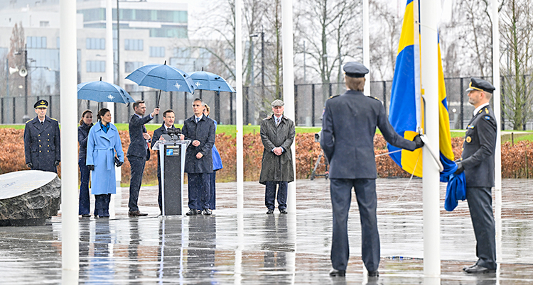 Det regnar. Ulf Kristersson håller ett tal utomhus. Två män ska hissa en svensk flagga.