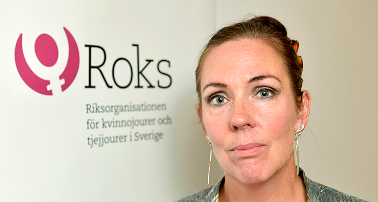 Nära bild på hennes ansikte. Bakom henne text på en vägg: Roks, Riksorganisationen för kvinnojourer och tjejjourer i Sverige, står det.