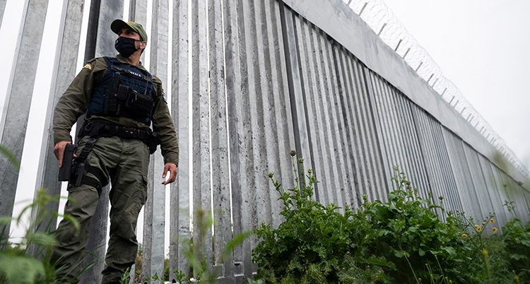 En polis står framför en hög mur. Längst upp på muren är det taggtråd.