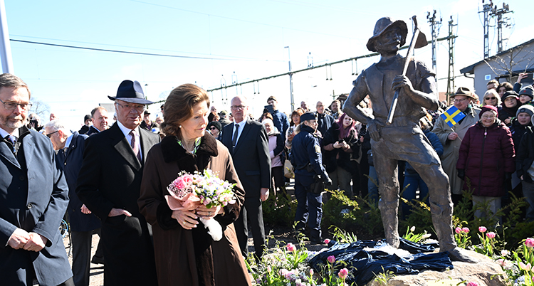 Kungen och drottningen står intill en staty. Bakom syns många människor.