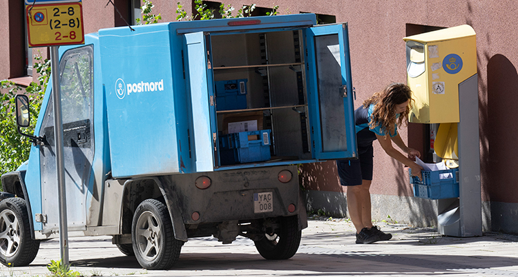 En brevbärare öppnar en gul brevlåda och håller en back under. Intill står en blå postbil.