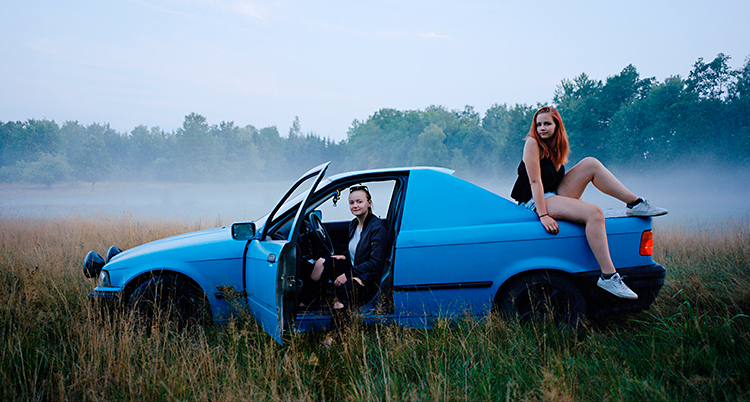 En blå epa står på ett fält. En tjej sitter bakom ratten. En annan tjej sitter bak på bilen.