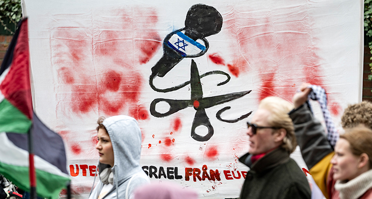 Människor står vid en banderoll med en bild på en sax som klipper av en mikrofon med Israels flagga på.