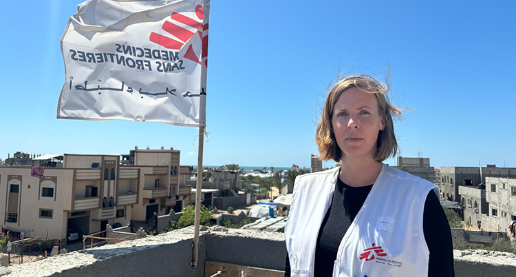 Hon har en vit väst på sig Hon står vid en flagga för organisationen Läkare utan gränser.