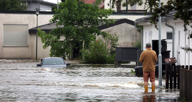 En man står och ser på en översvämmad gata, översvämmade hus och en bil som står i vattnet på gatan.