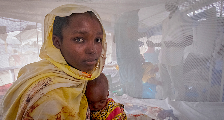 En kvinna sitter på en säng i ett läger för flyktingar. Hon håller i ett litet barn. Hon tittar in i kameran.
