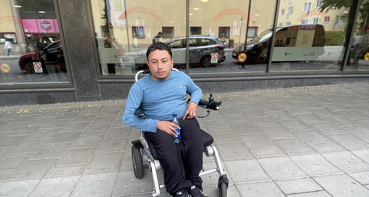 Han sitter i en rullstol på trottoaren utanför ett kontor.
