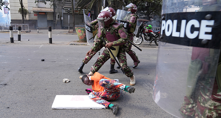 En person ligger på marken och försöker skydda sig. Poliser med sköldar och hjälmar slår mot honom.